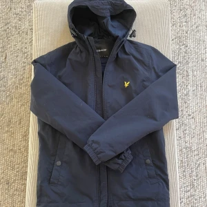 Jacka från Lyle & Scott - Tunn mörkblå skaljacka med huva från LYLE & SCOTT. Stängs med dragkedja fram och är åtdragbar i nederkant samt kring huva. Snedställda fickor med knapp i sidorna samt en innerficka. Foder i mesh och liten varumärkeslogo på vänster sida bröst.