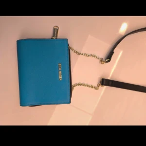 Blå axelväska från Steve Madden - Snygg blå axelväska från Steve Madden med guldfärgade detaljer och kedja. Väskan har en dragkedja och flera fack inuti för kort och småsaker. Perfekt för att bära det viktigaste med stil.