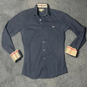 Marinblå skjorta från Burberry - Snygg blå skjorta från Burberry i slim fit. Skjortan har långa ärmar och klassiska Burberry-detaljer på insidan av kragen och ärmsluten. Perfekt för en stilren look. Skjortan ör köpt på sicuro fashion men passade inte. Den passar någon som är runt 170. Kontakta mig vid frågor eller funderingar🍻