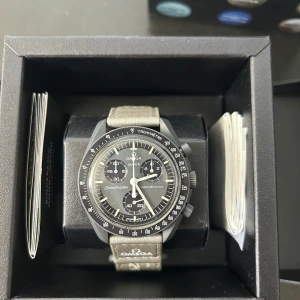 Omega moon swatch - Säljer en stilren Omega Speedmaster klocka med grått armband och svart urtavla. Klockan har en tachymeter och flera sub-dials för extra funktionalitet. Perfekt för den som uppskattar klassisk design och precision.