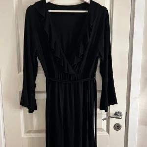 Svart långklänning med volangdetaljer - Elegant svart långklänning med volangdetaljer längs den v-ringade halsen. Klänningen har långa ärmar och ett knytband i midjan för en smickrande passform. Perfekt för en stilren och sofistikerad look.