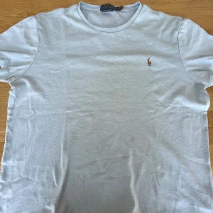 Ljus blå T-shirt från Polo Ralph Lauren - Säljer en stilren vit t-shirt från Polo Ralph Lauren med det klassiska broderade logotypen på bröstet. T-shirten har en normal passform och är tillverkad i mjuk bomull. Perfekt för en avslappnad look.