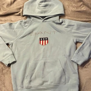 Grå hoodie från GANT - Säljer en stilren ljusblå hoodie från GANT med broderad logga och emblem på bröstet. Tröjan har en klassisk känguruficka och långa ärmar. Perfekt för en avslappnad stil.