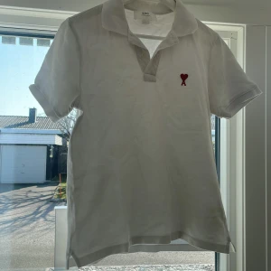 Vit kortärmad pikétröja från Ami - Säljer en stilren vit pikétröja från Ami med ett rött broderat hjärta och bokstaven A på bröstet. Tröjan har en klassisk krage och korta ärmar, perfekt för en avslappnad look.