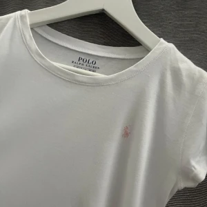 Vit t-shirt från Ralph Lauren - Säljer en klassisk vit t-shirt från Ralph Lauren med en liten rosa logga broderad på bröstet. T-shirten har en rund hals och korta ärmar. Perfekt för en stilren och enkel look. Passar till allt! 🌟