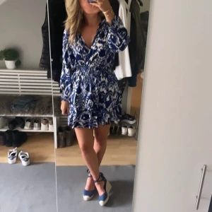 Blå och vit mönstrad klänning - Snygg kortklänning i blått och vitt med ett unikt mönster. Klänningen har långa ärmar och en v-ringad design som ger en elegant touch. Perfekt för sommarens alla tillfällen!