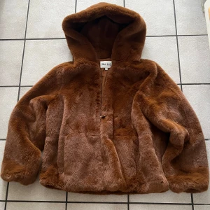 Brun pälsjacka från NA-KD - Säljer en supermjuk brun pälsjacka från NA-KD. Jackan har en mysig huva och är perfekt för kyliga dagar. Den är långärmad och har en stilren design utan synliga detaljer. Perfekt för att hålla sig varm och snygg i vinter! 🧥