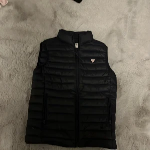 Svart dunväst från Guess - Snygg svart dunväst från Guess med dragkedja framtill och ett stilrent märke på bröstet. Perfekt för lager-på-lager under kyligare dagar. Västen har en modern design och är lätt att matcha med olika outfits. Dock två små hål ett på baksidan av västen som knappast syns och ett vid en av fickorna