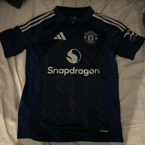 Manchester United bortaställ 24/25 fotbollströja  - Säljer en snygg mörkblå Manchester United fotbollströja från Adidas. Tröjan har korta ärmar och ett stilrent mönster. Den har vita detaljer och logotyper på bröstet och ärmarna. Perfekt för alla United-fans! 💙