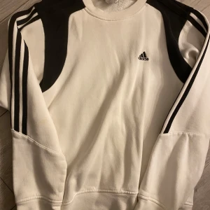 Vit och svart tröja från Adidas - Säljer en vit och svart tröja från Adidas med klassiska tre ränder längs ärmarna. Tröjan har en rund hals och långa ärmar. Perfekt för en sportig look eller avslappnad stil. Adidas-loggan är broderad på bröstet. Liten defekt vid ärmen vet ej om den går att få bort, strl xs