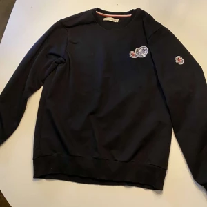 Svart tröja från Moncler - Säljer en stilren svart tröja från Moncler med rund hals och långa ärmar. Tröjan har Moncler-loggor på bröstet och ärmen. Perfekt för en casual look.
