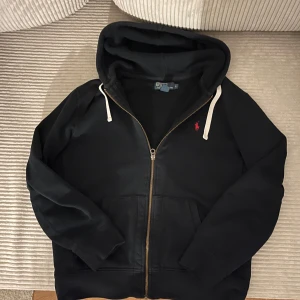 Mörkblå zip från Polo Ralph Lauren!sitter som en M! - Säljer en stilren svart hoodie från Polo Ralph Lauren med dragkedja och röd logga på bröstet. Tröjan har långa ärmar och en justerbar huva med vita snören. Perfekt för en avslappnad look.perfekt nu för våren och priset går att pruta! Vid frågor skriv 💬!