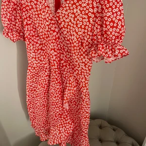 Röd blommig klänning från Chiquelle - Säljer en söt röd klänning med vita blommor från Chiquelle. Klänningen har korta ärmar med volangdetaljer och en v-ringad hals. Perfekt för midsommar eller annat tillfälle! Har sytt ihop v- ringningen lite men går att sprätta upp! Använd en gång 