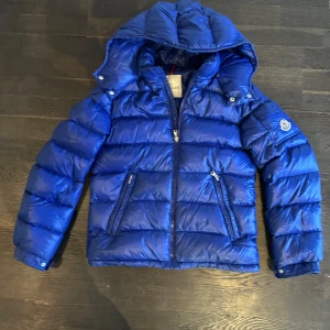 Blå dunjacka från Moncler - Säljer en snygg blå dunjacka från Moncler med huva och dragkedja. Jackan har långa ärmar och två fickor med dragkedja framtill. Perfekt för kyliga dagar.stl14/164
