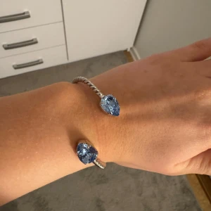 Silverfärgat armband med blå stenar - Snyggt silverfärgat armband med två stora blå stenar i ändarna. Armbandet har en vriden design som ger det en unik look. Perfekt för att lägga till en touch av färg och stil till din outfit.