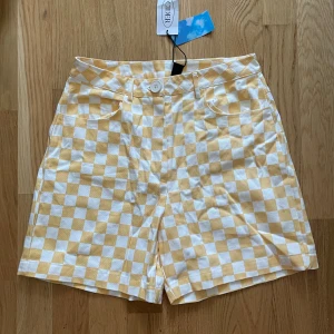 Gul- och vitrutiga shorts från Cider - Snygga gul- och vitrutiga shorts från Cider med hög midja och knappstängning. Perfekta för en solig dag med sitt fräscha rutmönster och bekväma passform. De har praktiska fickor både fram och bak. Aldrig använda, lapp kvar. Midjemått: ca 33cm. 100% cotton 