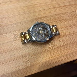 Klocka från Regal med länkarmband - Snygg klocka från Regal med en kombination av silver och guld på länkarmbandet. Urtavlan har romerska siffror och tre mindre urtavlor för extra funktioner. Boetten är i rostfritt stål och klockan är vattenresistent. Perfekt för den som gillar en klassisk och stilren look.