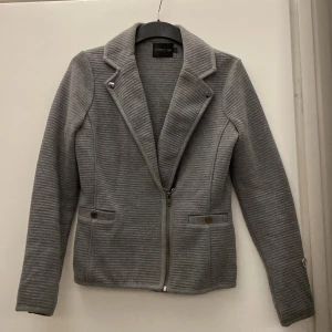 Grå jacka från Stockh LM - Snygg grå jacka från Stockholm med dragkedja framtill och stilrena knappar på kragen. Jackan har en ribbad textur och långa ärmar, perfekt för en chic look. Den är i en normal passform och passar bra till både vardag och lite finare tillfällen.