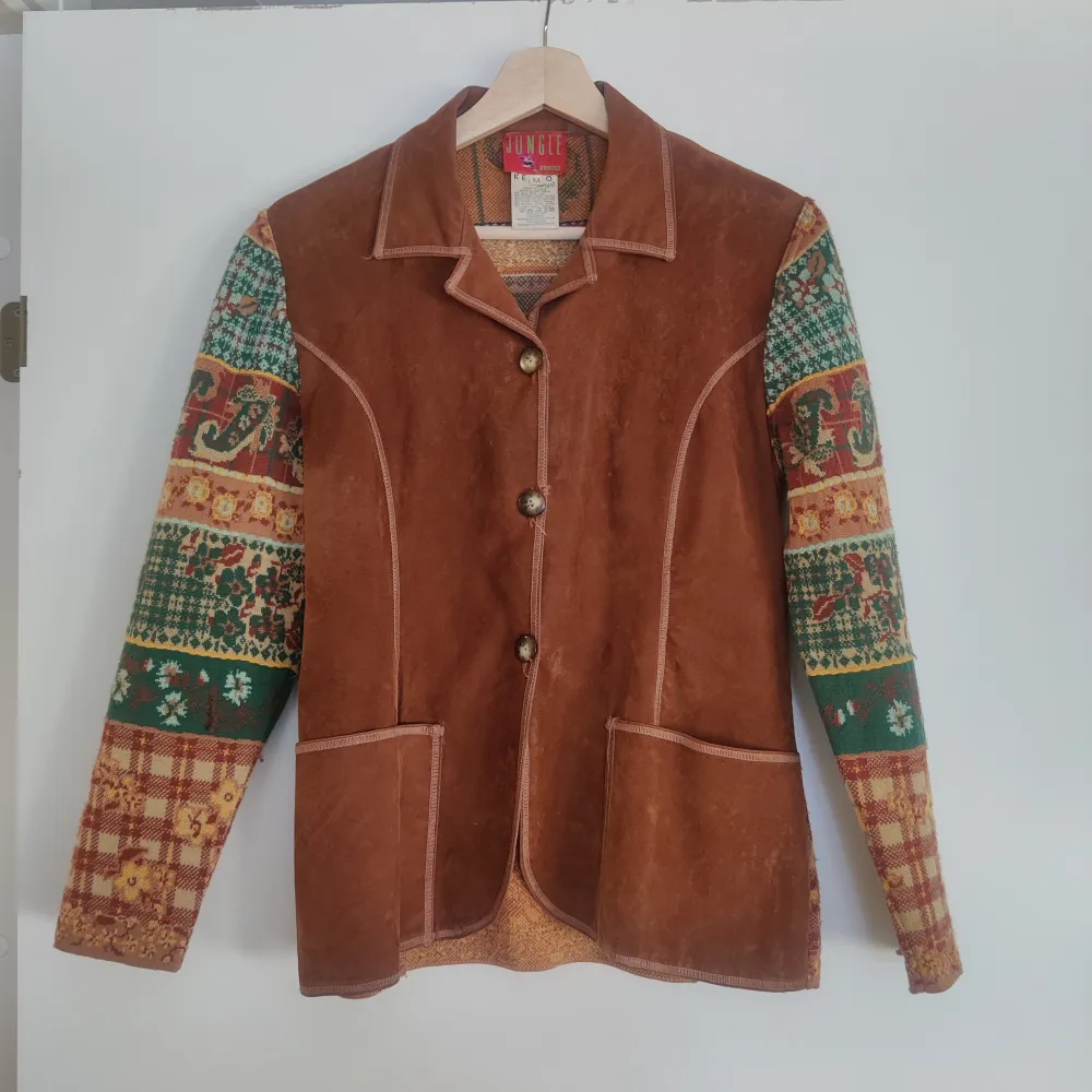 Unik brun mockajacka från Kenzo med färgglada mönstrade ärmar i grönt, rött och gult. Jackan har knappar framtill och två fickor. Perfekt för dig som vill sticka ut med en stilfull och annorlunda look.. Takit.
