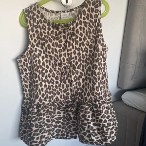 Leopardmönstrad ärmlös topp från ZARA - Säljer en snygg leopardmönstrad ärmlös topp från ZARA. Toppen har en söt rosettdetalj framtill och en volangkant nedtill som ger en feminin touch. Perfekt för att liva upp garderoben med lite djurmönster!