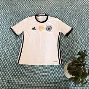 Vit fotbollströja från Adidas - Säljer en vit fotbollströja från Adidas med svart krage och detaljer. Tröjan har korta ärmar och ett tryck av Deutscher Fussball-Bund-loggan på bröstet. Perfekt för fotbollsfans! ⚽⚠️står att varan är i 164cm men känns som 146-158cm