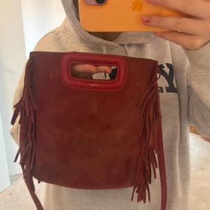 Röd axelväska med fransar från Maje - Snygg röd axelväska från Maje i mocka med coola fransdetaljer. Väskan har ett praktiskt handtag och en justerbar axelrem. Perfekt för att ge din outfit en bohemisk touch.