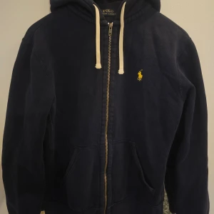 Mörkblå hoodie från Ralph Lauren - Säljer en snygg mörkblå hoodie från Ralph Lauren med dragkedja och huva. Den har en broderad gul logga på bröstet och vita snören vid huvan. Perfekt för en avslappnad stil.