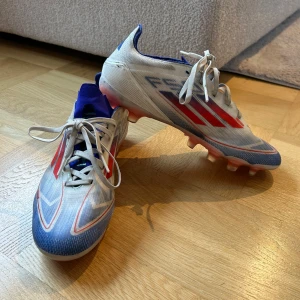 Adidas F50 Pro MG fotbollsskor - Snygga Adidas F50 Pro MG fotbollsskor i vitt med röda och blå detaljer. Skorna har snörning och mg dobbar som är multi grass och passar för både gräs och konstgräs. Ny pris är 1700 kr från unisport. Skorna är i mycket bra skick då dem bara är använda ett fåtal gånger.