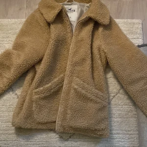 Beige teddyjacka från Hollister - Mysig beige teddyjacka från Hollister med långa ärmar och två stora fickor framtill. Perfekt för kyliga dagar när du vill hålla dig varm och stilren. Jackan har en klassisk krage och stängs med dragkedja. Helt ny men för stor