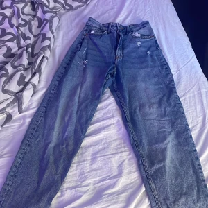 Blå jeans med slitningar - Säljer ett par blå jeans med slitningar på framsidan. De har en klassisk femficksdesign och en dragkedja med knappstängning. Perfekta för en avslappnad stil.