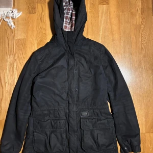 Svart jacka från Peak Performance - Säljer en svart jacka från Peak Performance med rutigt foder i rött och vitt. Jackan har dragkedja och knappar framtill samt stora fickor. Perfekt för kyligare dagar med sin stilrena design och praktiska detaljer.