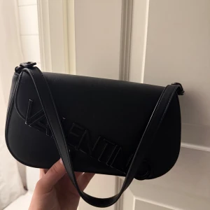 Svart axelväska från Valentino - Snygg svart axelväska från Valentino med stilren design. Väskan har en elegant axelrem och är perfekt för att bära dina nödvändigheter. Den har en subtil logotypdetalj på framsidan som ger en exklusiv touch.