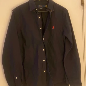 Mörkblå skjorta från Ralph Lauren - Säljer en stilren mörkblå skjorta från Ralph Lauren i slim fit. Skjortan har långa ärmar och knappar framtill. Den är dekorerad med det klassiska röda broderade logotypen på bröstet. Perfekt för en elegant look. 1200kr nypris.