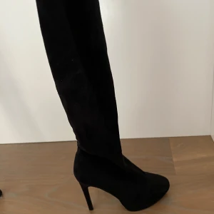 Svarta overknee boots med klack - Snygga svarta overknee boots från Nelly med hög klack och dragkedja på sidan. Skorna är i mocka och har en spetsig tå. Använda en gång så sulan är i mycket bra skick och finns 4 extra ”klackar” man kan byta när de blir slitna. 