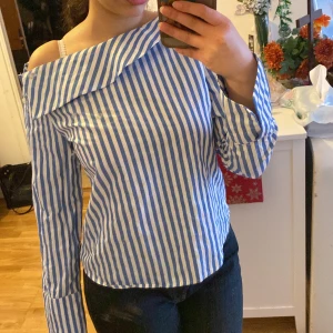 Blåvit randig offshoulder blus från Gina Tricot - Säljer en snygg blåvit randig offshoulder blus från Gina Tricot. Blusen har långa ärmar och en unik design med knappar på sidan. 