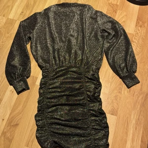 Glittrig klänning från H&M Divided - Snygg glittrig klänning från H&M Divided med lång ärm och rynkad detalj i midjan. Perfekt för en kväll ute med vänner. Klänningen har en tight passform och är i en mörk färgton.