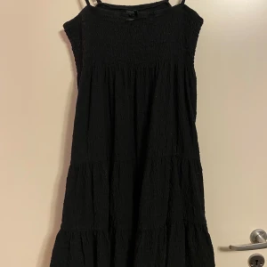Gina Tricot svart klänning, storlek L - Svart klänning med smala axelband och smock överdel. Klänningen har en söt liten rosett framtill och en vågig nederkant. Strl L men liten i strl, passar strl M.