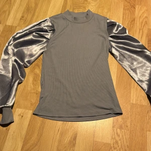 Ljusblå topp med glansiga ärmar från Shein - Säljer en snygg ljusblå topp från Shein med glansiga, långärmade ärmar. Toppen har en ribbad struktur och en hög krage. Perfekt för att ge en stilren look med en touch av glamour.