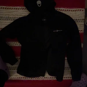 Svart jacka från Sail Racing - Snygg svart jacka från Sail Racing med dragkedja och huva. Perfekt för en stilren look. Jackan har långa ärmar och en diskret logga på bröstet.