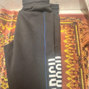 Hoodrich byxor - Säljer ett par svarta mjukisbyxor med vit text längs benet och en blå linje som detalj. Perfekta för en avslappnad stil.