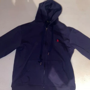 Mörkblå hoodie från Ralph Lauren - Säljer en snygg mörkblå hoodie från Ralph Lauren med dragkedja och huva. Den har en liten röd logga på bröstet och snörning vid huvan. Perfekt för en casual look! Köpt från kidsbrandstorr! säljer pågrund av att jag råkade köpa fel storlek