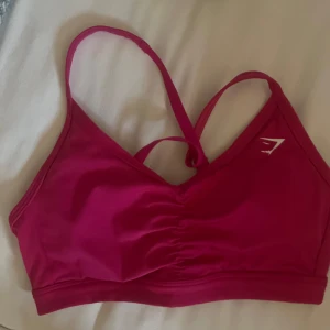 Rosa sporttopp från Gymshark - Säljer en snygg rosa sporttopp från Gymshark med smala axelband och korsad rygg. Perfekt för träning och ger bra stöd. Toppen har en enkel design med en liten logga framtill. Passar perfekt för gymmet eller yoga.