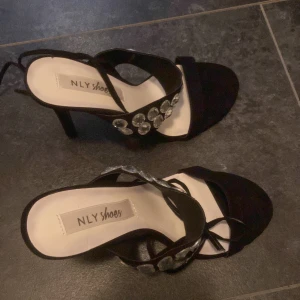 Svarta klackskor från NLY Shoes - Eleganta svarta klackskor från NLY Shoes med dekorativa stenar på remmen. Skorna har en öppen tå och knytning runt ankeln för en stilren look.