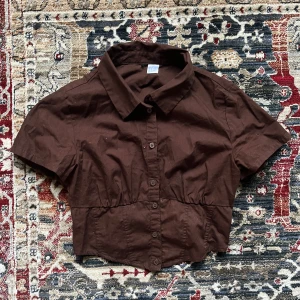 Brun croppad skjorta! - Säljer en brun croppad skjorta med korta ärmar och knappar framtill. I storlek S och har en tight passform vilket gör att den sitter snyggt mot kroppen!  Skjortan har en elastisk detalj i ryggen för en bekväm passform. Perfekt för en stilren look! 🩷