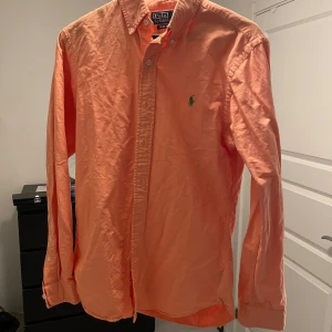 Orange skjorta från Ralph Lauren - Säljer en snygg orange skjorta från Ralph Lauren i slim fit. Skjortan har långa ärmar och knappar framtill samt en broderad logga på bröstet. Perfekt för en stilren look inför sommaren.