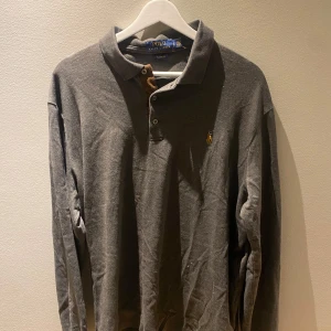 Grå pikétröja från Ralph Lauren - Säljer en klassisk grå pikétröja från Ralph Lauren med broderad logotyp på bröstet. Tröjan har långa ärmar och en knappslå framtill. Perfekt för en stilren look.