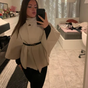 Beige kappa med bälte - Snygg beige kappa med bälte i midjan och vida ärmar. Perfekt för en stilren look. Kappan har en elegant design med en knappdetalj vid halsen.