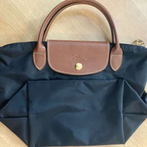 Svart handväska från Longchamp - Snygg svart handväska från Longchamp med bruna skinnhandtag och detaljer. Väskan har en praktisk dragkedja och en knapp framtill. Perfekt för att bära dina dagliga nödvändigheter med stil.