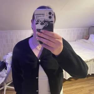 Svart kofta från Kappahl med rosett - Säljer en elegant svart kofta från Kappahl med dekorativa knappar och en stor rosett framtill. Perfekt för att ge en stilren touch till din outfit. Koftan har långa ärmar och är tillverkad i ett mjukt material.