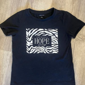 Svart t-shirt med zebramönster från Ellos - Snygg svart t-shirt från Ellos med ett coolt zebramönster och texten 'Always believe in hope yourself' på framsidan. Perfekt för en avslappnad stil.